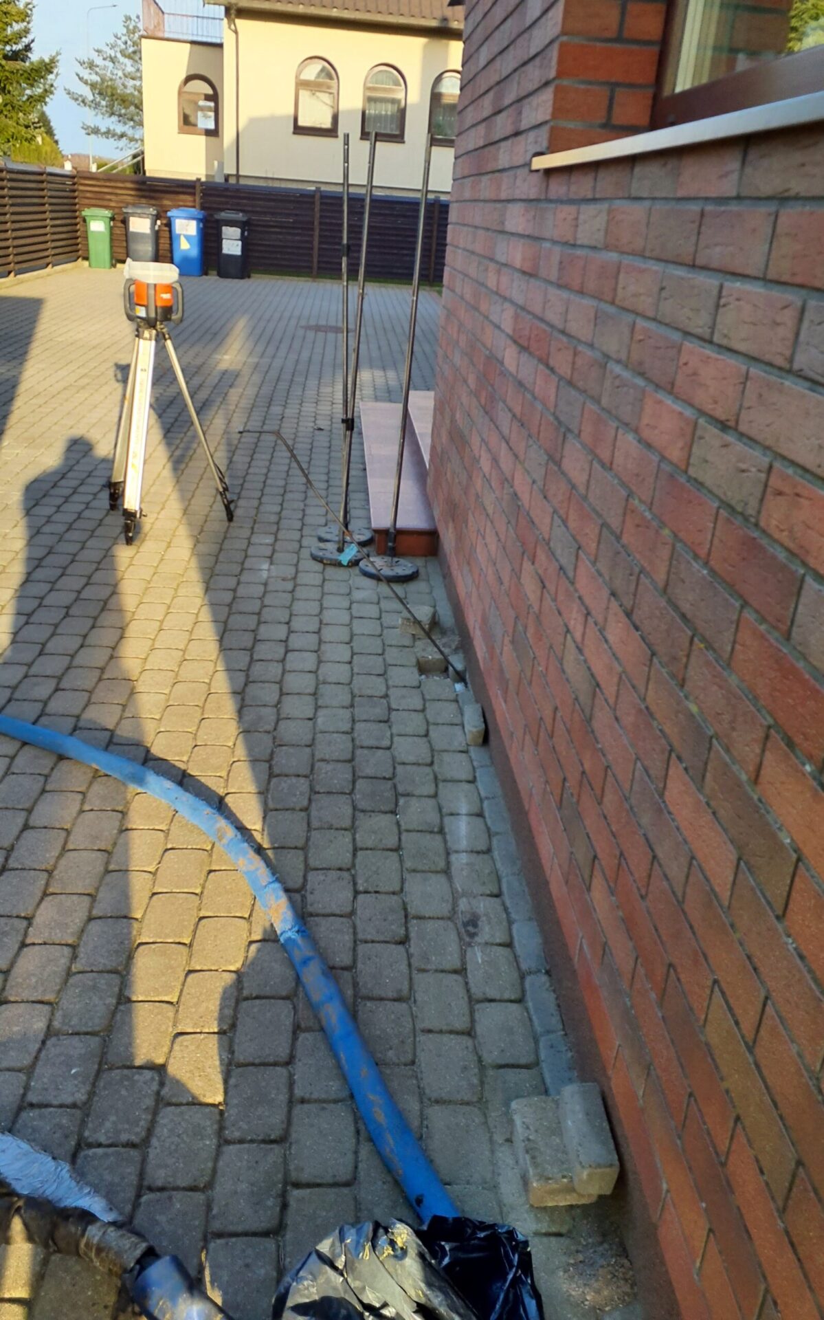 Wąż iniekcyjny, niwelator na statywie i łaty pomiarowe przy ścianie domu z cegły, kostka brukowa wokół budynku pozostaje na miejscu
