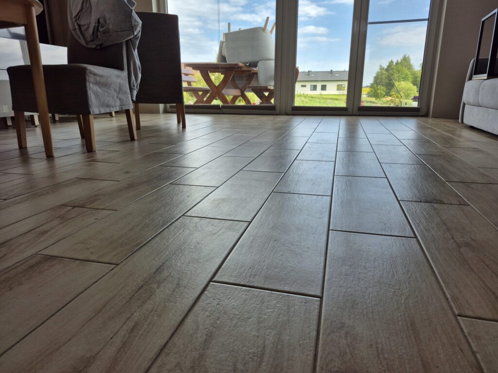 Podłoga z płytek drewnopodobnych w salonie widziana z poziomu posadzki, w tle okna tarasowe